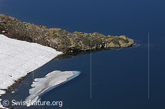 Foto: Halbinsel in dunkelblauem Bergsee. In der Nähe des Ufers befindet sich eine Eisscholle.