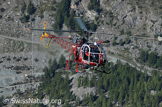 Foto: Helikopter im Gebirge. Der Pilot steuert die Maschine mit der am Seil hängenden Last über eine Gebirgsbaustelle.