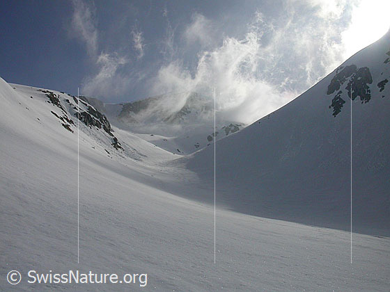 Foto: Sturm mit Wolkenfetzen und Schneefahnen über dem obersten Teil des Hotäl im Gegenlicht. Die Aufnahme entstand am frühen Morgen.