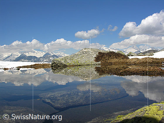 Foto: Blick über den Abfluss des Bergsees. Im Wasser sind Felsblöcke zu sehen. Tolle Spiegelung mit Felsblock, Schnee, den Berner Alpen, Schönwetterwolken und blauem Himmel.