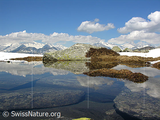 Foto: Blick über den Abfluss des Bergsees. Im Wasser sind Felsblöcke zu sehen. Tolle Spiegelung mit Felsblock, Schnee, den Berner Alpen, Schönwetterwolken und blauem Himmel.