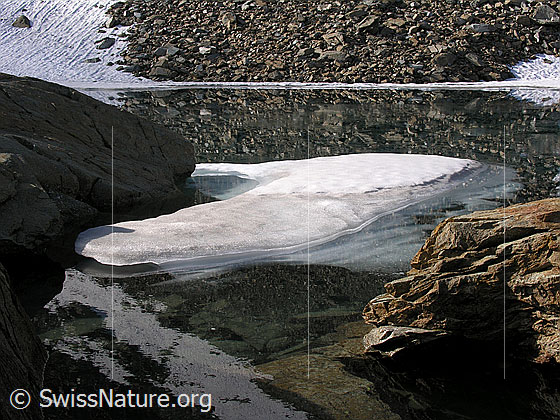 Foto: Spiegelung einer Eisscholle und einer Geröllhalde im Zantmärjelesee. Im Vordergrund ist ein rötlicher Felsblock zu sehen.