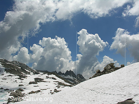 Foto: Blick über ein Schneefeld mit Spur Richtung gezacktem Felsgrat im Horizont. Dahinter türmen sich mächtige Wolken.
