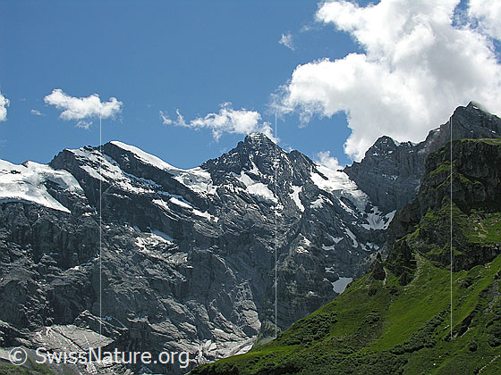 Foto: Tischingelspitz und Gspaltenhorn mit Quellwolken am blauen Himmel und Sonnenlicht auf den steil abfallenden Felswänden.