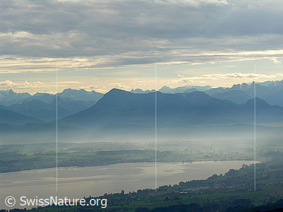 Foto: Luftaufnahme Berge und See mit Wolkenstimmung. Rigi mit feiner Nebelschicht über dem See und Bergkette im Hintergrund.