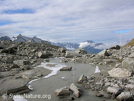 Foto: Blick über einen ruhigen Bergbach mit gefrorenem Ufer, welcher durch Geröll und Felsblöcke fliesst. Im Hintergrund sind die Schinhörner und das Bättlihorn zu sehen.