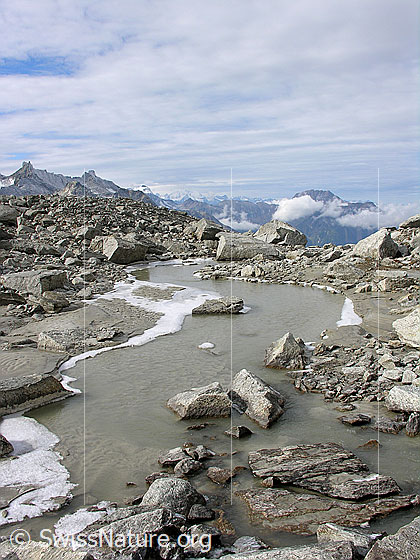 Foto: Blick über einen ruhigen Bergbach mit gefrorenem Ufer, welcher durch Geröll und Felsblöcke fliesst. Im Hintergrund sind die Schinhörner und das Bättlihorn zu sehen.