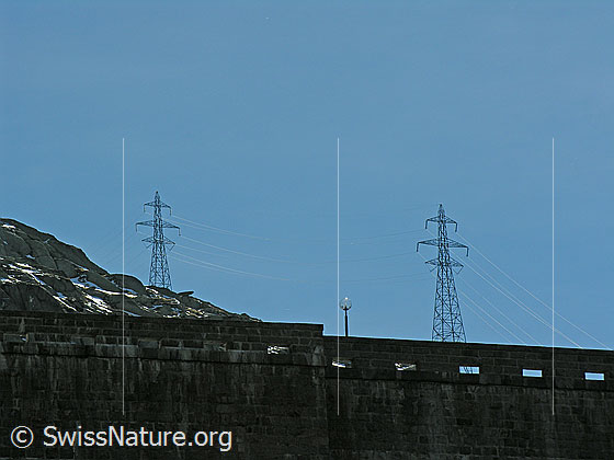 Foto: Blick über die Krone der Staumauer Seeuferegg (Grimselsee) auf zwei Masten einer Hochspannungsleitung.