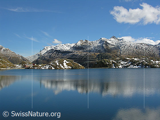 Foto: Blickvom Grimselpass über den Totesee Richtung Süden. Eine Bergkette mit verschneiten Gipfeln und eine Wolke spiegeln sich im blauen Wasser.