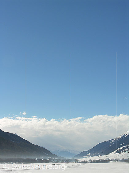 Foto: Blick von Obergesteln Richtung Ulrichen. Auf der Loipe sind einige Personen unterwegs. Aus Westen nähert sich eine Front und bringt dichte Wolken.