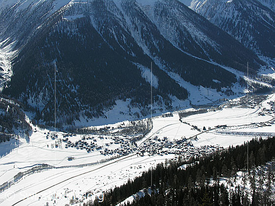 Foto: Tiefblick in den Talgrund des Obergoms. Zu sehen sind das Dorf Reckingen und steile Berghänge mit Schutzwäldern.