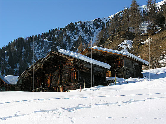 Foto: Alte Oberwalliser Häuser in Winterlandschaft und Spuren im Schnee. Im Hintergrund ist eine steile Bergflanke zu sehen.