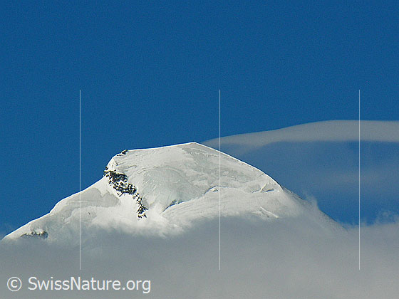 Foto: Der Gipfel des Allalinhorns ragt aus einer Wolkenschicht. Darüber blauer Himmel mit einer Schleierwolke. Das Allalinhorn ist einer der einfachsten Viertausender der Schweiz. Entsprechend oft wird das Allalinhorn bestiegen.