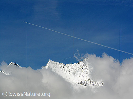 Foto: Durch Wolken sind Dom, Nadelhorn und Lenzspitze zu sehen. 
Darüber ein Kondensationsstreifen eines Flugzeugs.