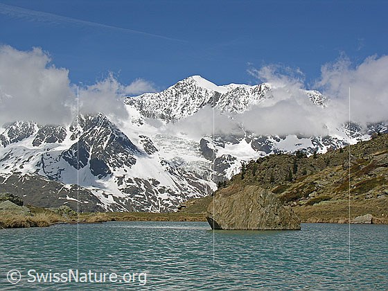 Foto: Balfrin mit Wolken. Davor ein Bergsee (Farbe: türkis). Im Bergsee ein Felsblock.
