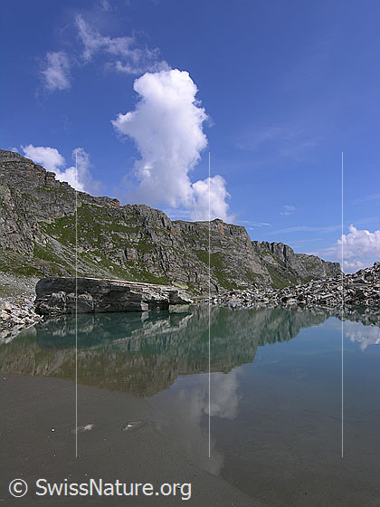 Foto: Blick über den Mättitalsee in welchem sich ein grosser Felsblock und ein Grat spiegeln. Im Hintergrund steigt eine weisse Wolke auf.
