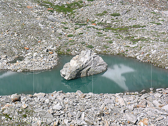 Foto: Blick auf den Mättitalsee mit grossem Felsblock in der Mitte des türkisfarbenen Bergsees. Der See ist von zahlreichen Steinen und Felsblöcken umgeben. Der grosse Felsblock im Mättitalsee glitt im Winter 2005/2006 über den Schnee direkt in den See.