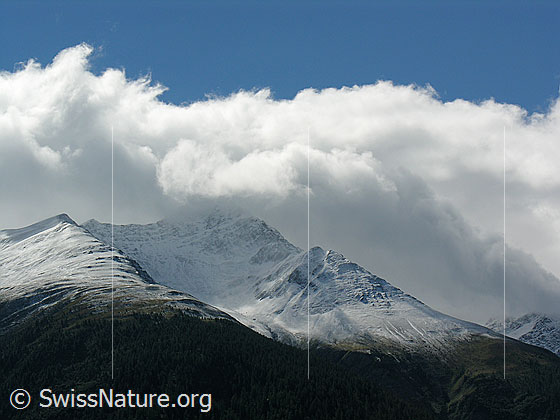 Foto: Deutlich sichtbare Schneefallgrenze nach einem kurzen Wintereinbruch. Der Gipfelbereich des Bättlihorns ist mit Neuschnee überzuckert. Darüber türmen sich Wolken und es fällt Schatten auf die Berglandchaft.