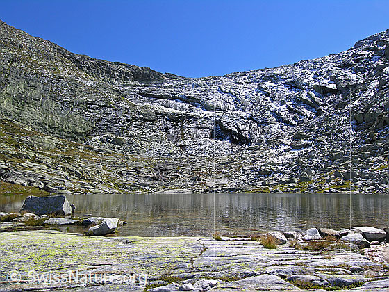 Foto: Witentirsee und leicht mit Schnee überzuckerte Felsstufe zum Witentir. Im Vordergrund: Vom Wasser geschliffener Fels.