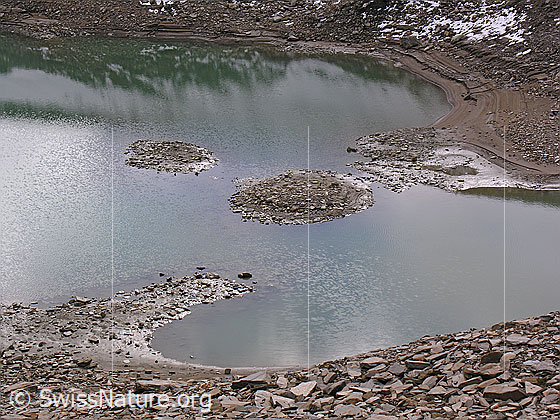 Foto: Bergsee bei niedrigem Wasserstand. Kleine Inseln kommen zum Vorschein. Das Ufer besteht aus Geröll, Sand und Morast. Durch die stufenweise Absenkung des Wasserspiegels haben sich im Sand interessante Strukturen gebildet.