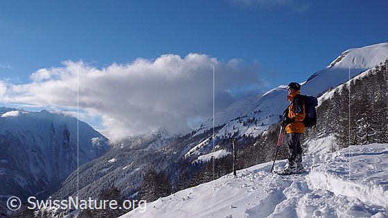 Foto: Schneeschuhläuferin blickt über das verschneite Binntal. Am blauen Himmel über dem Bergtal ist eine grosse Wolke zu sehen.