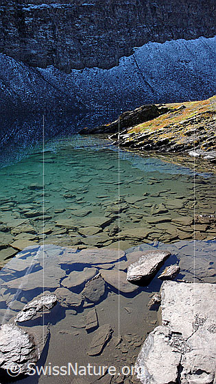 Foto: Hagelseeli mit Spiegelung von Schneeresten am Fusse einer Felswand. Durch das klare, türkisfarbene Wasser sind Steinplatten zu sehen.
