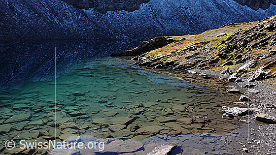 Foto: Hagelseeli mit Spiegelung von Schneeresten am Fusse einer Felswand. Durch das klare, türkisfarbene Wasser sind Steinplatten zu sehen.