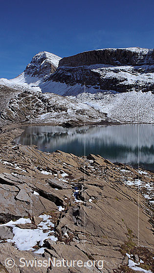 Foto: Häxeseeli mit schwacher Spiegelung. Die Wart, das Schwarzhorn und die Grosse Chrinne sind leicht verschneit. Der Bergsee ist von bräunlichen Felsen umgeben.
