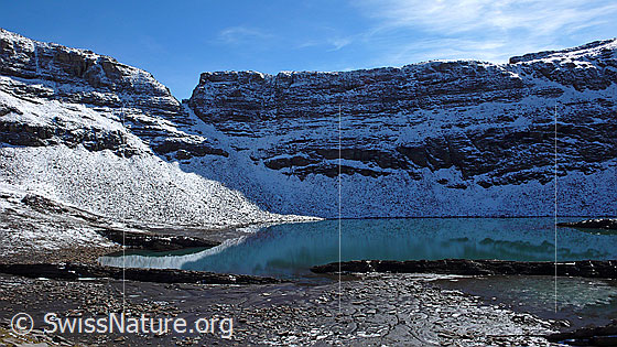 Foto: Häxeseeli (Grindelwald) mit seichtem, lehmigem Ufer. Der Bergsee liegt grösstenteils im Schatten. Dahinter die leicht verschneite Kleine Chrinne mit fein gezeichneten Felsbändern.