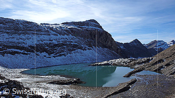 Foto: Häxeseeli (Grindelwald) mit schwacher Spiegelung und seichtem Ufer. Der Bergsee liegt grösstenteils im Schatten. Felsschichtungen ragen aus dem Wasser und bilden die Begrenzung des Hochtals Hinterbirg. Dahinter die leicht verschneite Kleine Chrinne mit fein gezeichneten Felsbändern sowie die Grossenegg.