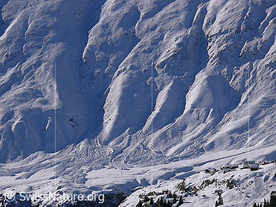 Foto: Lawinenkegel am Fuss einer gefurchten Bergflanke. Auf dem Bergrücken im Vordergrund ist ein Bergdorf in der sonnigen Winterlandschaft zu sehen.