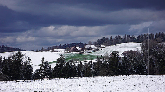 Foto: Wolkenstimmung über Winterlandschaft im Emmental. Zu sehen sind ein Weiler mit Bauernhöfen umgeben von schneebedeckten Hügeln und Wald. In einem Feld schimmert das Grün einer Grasfläche durch die schwindende Schneedecke.