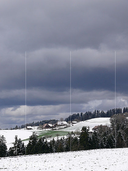 Foto: Wolkenstimmung über Winterlandschaft im Emmental. Zu sehen sind ein Weiler mit Bauernhöfen umgeben von schneebedeckten Hügeln und Wald. In einem Feld schimmert das Grün einer Grasfläche durch die schwindende Schneedecke.
