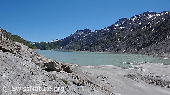 Foto: Oberaarsee mit Sandstrand und Felsblöcken auf abgeschliffenen Felsen im Uferbereich. Im Hintergrund sind Sidelhorn und Gross Sidelhorn zu sehen.