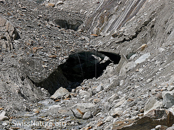 Foto: Eisbrücke am Gletscherrand. Ein kleiner Bach mit Gletscherwasser fliesst zwischen Geröll hindurch. Von der Eisbrücke rinnt Schmelzwasser. Instabiles Gelände zufolge Gletscherrückzug und Erosion.