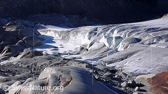 Foto: Gefrorener Gletschersee an der Gletscherzunge des Rhonegletschers. Im Vordergrund vom Gletscher abgeschliffene Felsen (Gletscherschliff).
