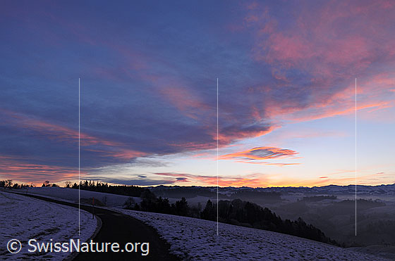 Foto: Wolkenfeld und Morgenrot über der Hügellandschaft des Emmentals. Ein Fahrweg führt durch die schneebedeckte Landschaft.
