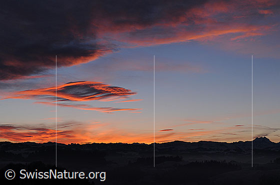 Foto: Wolkenfelder in kräftigem Morgenrot und Silhouette einer Bergkette. Die Wetterlage (Föhn) hat zur Entstehung der interessanten Wolkenformen beigesteuert.
