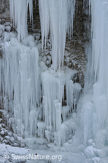 Foto: Leicht verschneite Eiskaskade an Felswand. Die einzelnen Eiszapfen sind zu einem Eisfall mit gerundeten und spitzen Formen verschmolzen.