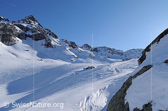 Foto: Schneebedeckte Berglandschaft mit Ofenhorn, langen Schattenwürfen, Abfahrtsspuren und teilweise zugewehter Aufstiegsspur. Im Vordergrund eine Schneeverwehung.