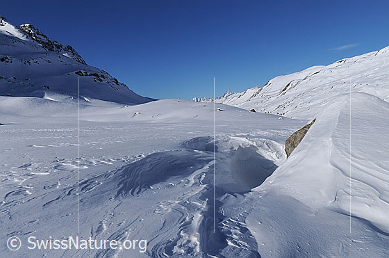 Foto: Schneeverwehung um einen Felsblock in sonniger Berglandschaft.