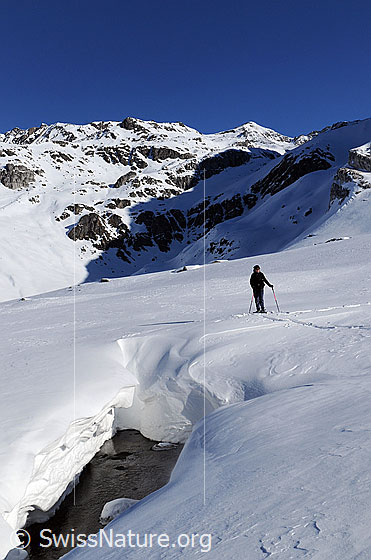 Foto: Schneeverwehung an offener Stelle eines Bergbachs. Am verwächteten Ufer steht eine Schneeschuhläuferin. Im Hintergrund ist das Hohsandhorn zu sehen.