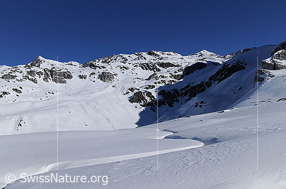 Foto: Schneebedeckte Binntaler Berglandschaft mit Turbhorn, Strahlgrät und Hohsandhorn. Durch die Schneefläche im Vordergrund verläuft ein zugeschneiter Bergbach.