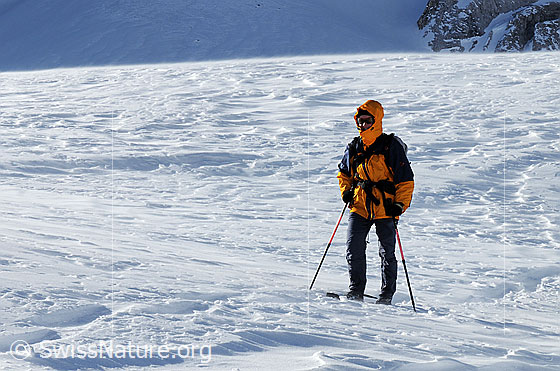 Foto: Schneeschuhläuferin im Föhnsturm. Die Schneeoberfläche weist schöne Strukturen der Verwehungen auf. Der Föhn blies an diesem Tag stark über den nahen Albrunpass. Das Gelände wirkte wie eine Düse, welche den Wind beschleunigt. Ca. 100 Höhenmeter tiefer war es nahezu windstill.