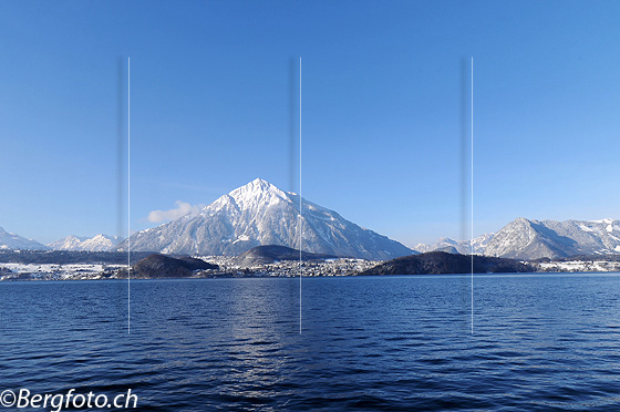 Foto: Blauer See und verschneiter Berg. Blick über die leicht gewellte Wasserfläche des Thunersee zum Niesen mit Neuschnee.