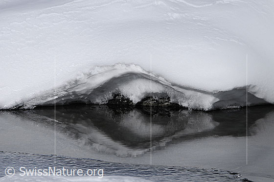 Foto: Spiegelung der Schneedecke und der Steine am Ufer im teilweise gefrorenen Wasser.
