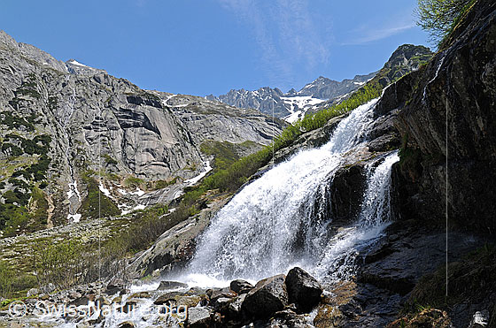 Foto: Frisch sprudelnder Wasserfall umgeben von grünen Kleinsträuchern, hohen Felswänden und den Hinteren Gelmerhörnern.