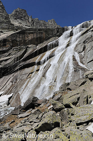 Foto: Hoher Wasserfall mit mehreren Armen und Verzweigungen ergiesst sich über eine plattige Felswand. Im Horizont sind die Gelmerhörner zu sehen.