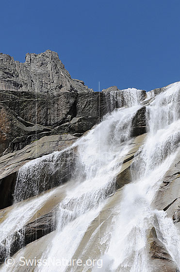 Foto: Breiter Wasserfall fällt über glatte, geneigte Felsplatten. Im Hintergrund ragt ein Felsturm des Klettergebiets Gelmerhörner in die Höhe.