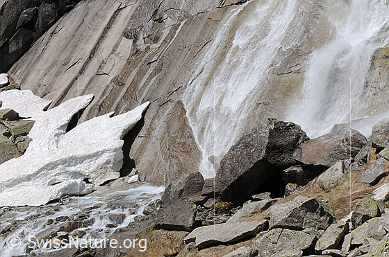 Foto: Schneereste und Wasserfall am Fuss einer Felswand. Das Wasser fällt über die glatten Felsen und fliesst als Bergbach weiter.